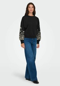 Črna trenirka z cvetličnimi okraski na napihnjenih rokavih, v kombinaciji s širokimi modrimi traper hlačami in črnimi gležnjarji. Gladek material, sproščen kroj.