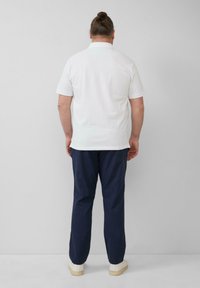 Mann mit braunen Haaren zu einem Dutt gebunden, trägt ein weißes Kurzarmhemd, eine marineblaue Hose und weiße Sneakers, steht mit dem Gesicht zu einer weißen Wand.