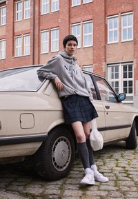 Pelēks Nike kapučjaka, tumši zila svārku kostīms ar rakstu, pelēkas ceļgala augstās zeķes, baltas kedas. Stāv blakus bēšai automašīnai ar netīru riteni uz akmens bruģa.