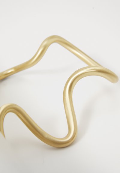 Glatt, guldfarvet metalarmbånd med et åbent, bølget design, der ligger på en hvid overflade.