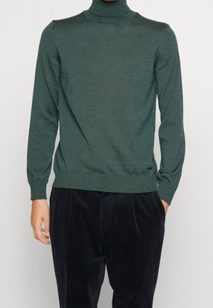 Mężczyzna w ciemnozielonym golfie i czarnych sztruksowych spodniach, z rękami częściowo w kieszeniach, na jednolitym tle.