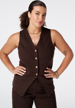Femme aux cheveux bouclés portant un gilet marron sans manches boutonné et un pantalon, accessoirisée avec des bijoux en or et un petit tatouage sur le bras, debout les mains sur les hanches.