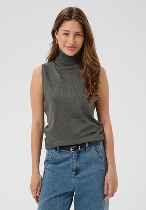 Jeune femme aux longs cheveux bruns portant un pull gris sans manches à col roulé et un jean bleu sur un fond uni.