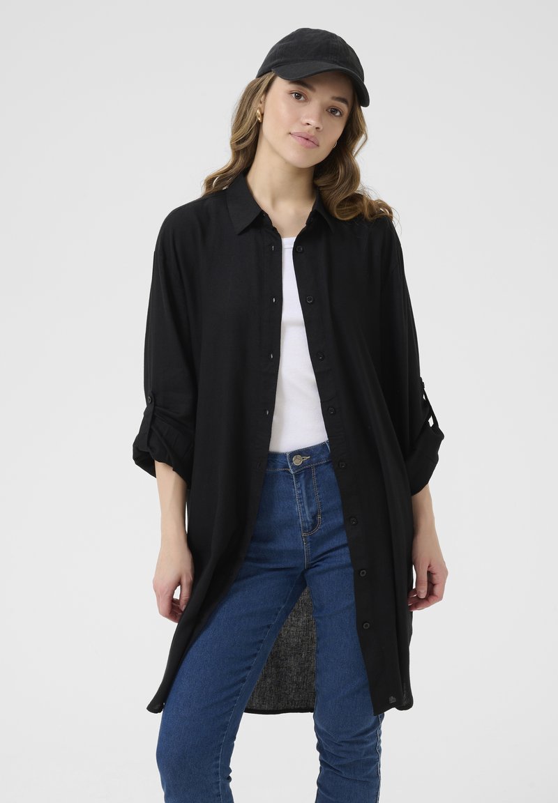 Svart oversized skjorte med knappelukking foran, rullede ermer og en lang, Rett kutt. Kombinert med blå jeans og en svart caps.