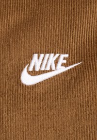 Brūns kordūru audums ar izšūtu balto Nike logotipu un swoosh dizainu, akcentējot ribotu tekstūru un izteiksmīgu marku.