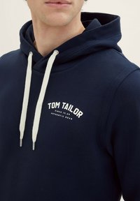 Felpa con cappuccio blu navy realizzata in tessuto morbido, con lacci bianchi e un logo stampato che recita "TOM TAILOR" sul petto.