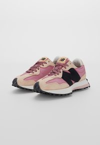 Pár růžových a béžových tenisek New Balance s černými akcenty loga a bílými podrážkami na jednobarevném světlém pozadí.