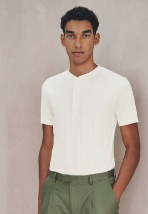 Joven con el pelo rizado que lleva una camisa blanca de manga corta abotonada metida dentro de pantalones verde oliva, de pie frente a un fondo liso.