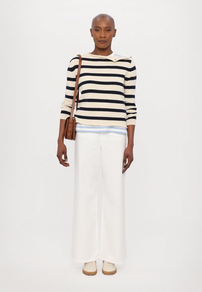 Maglione a righe in crema e blu navy, pantaloni bianchi a gamba larga, scarpe beige e una piccola borsa a tracolla marrone. Il maglione presenta dettagli con bottoni sulle spalle.