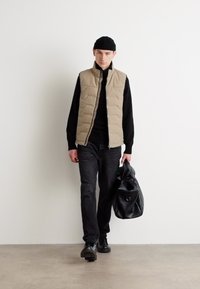 Beige puffer vest over en svart genser, svarte jeans og svarte sko. Modellen bærer en stor svart veske, stående mot en hvit bakgrunn.