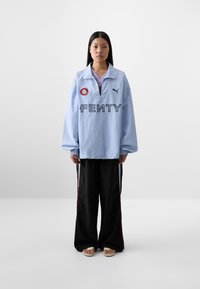 A young woman stands against a plain background, wearing an oversized light blue Fenty sweatshirt and black pants with side stripes.