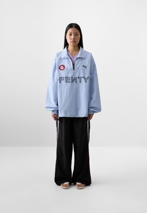 Una giovane donna è in piedi contro uno sfondo semplice, indossando una felpa Fenty oversize di colore azzurro chiaro e pantaloni neri con strisce laterali.
