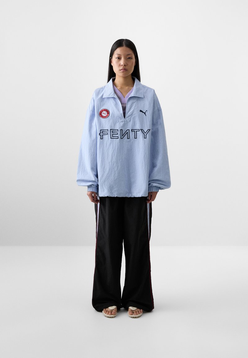 A young woman stands against a plain background, wearing an oversized light blue Fenty sweatshirt and black pants with side stripes.
