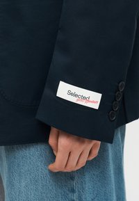 Κοντινό πλάνο ενός ατόμου που φοράει σκούρο σακάκι με τέσσερα κουμπιά στο μανίκι και μια λευκή ετικέτα που γράφει "Selected Hotel Amour."