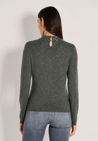 Frau mit langen, dunklen Haaren, die einen enganliegenden grauen Pullover mit einem mit Perlen besetzten hohen Kragen und einem Schlüssellochausschnitt trägt, kombiniert mit hellblauen Jeans.