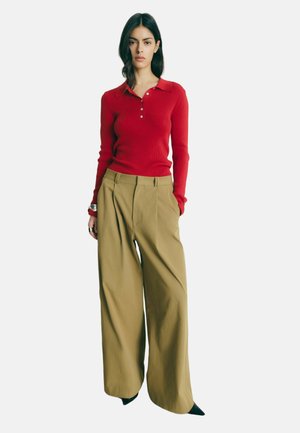 Kvinde med langt mørkt hår iført en rød, langærmet skjorte med krave og beige bredbenede bukser, stående med hænderne i lommerne.