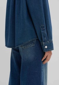 Camicia di denim di colore blu scuro, maniche lunghe e polsini a punta. Presenta un orlo curvo e un bottone a pressione argentato sul polsino.