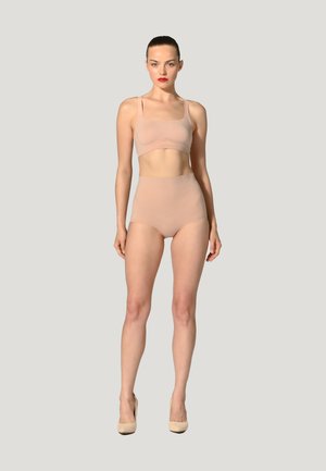 Vrouw staat, draagt naaktkleurig hooggetailleerd ondergoed en bijpassende crop top met naaktkleurige hoge hakken tegen een effen lichte achtergrond.