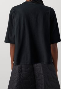 Musta puuvillainen lyhythihainen T-paita, jossa on rento istuvuus, pyöreä kaula-aukko ja suora helma. Takana on minimaalinen muotoilu.