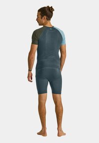 Man with brown hair in a bun wearing a two-tone short-sleeve shirt and matching shorts stands barefoot, turned away against a plain background.