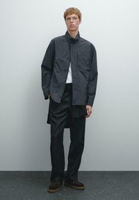 Giacca button-up grigio scuro con un alto colletto, indossata sopra una camicia bianca; abbinata a pantaloni neri larghi e scarpe in suede marroni.