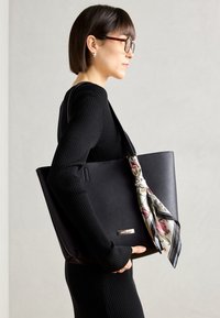 Bolso de mano de cuero negro con superficie suave, con un pañuelo de seda con estampado floral atado en el asa y un acento en oro del logo.