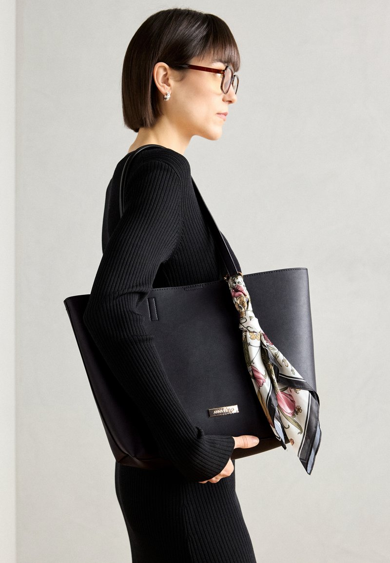Bolso de mano de cuero negro con superficie suave, con un pañuelo de seda con estampado floral atado en el asa y un acento en oro del logo.
