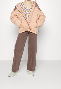 Ljusrosa puffjacka med bruna accenter, elastiska manschetter och dragsko vid nederkanten. Bärs över en mönstrad tröja och bruna vida byxor.