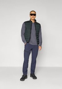 Juodas pūkuotas liemenė virš pilkos ilgomis rankovėmis palaidinės, derinama su tamsiai mėlynomis kelnėmis ir juodais batais; dėvimos saulės akiniai, stovintis prieš neutralią foną.