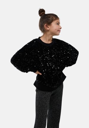 Sweat-shirt noir à sequins avec une coupe oversize et des poignets côtelés, associé à un pantalon gris scintillant. Fond blanc uni.