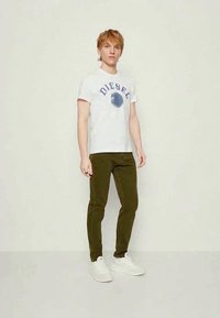 Fehér póló "DIESEL" felirattal, olívazöld, slim-fit nadrág és fehér sportcipő. A modell egyenesen áll egy semleges háttér előtt.