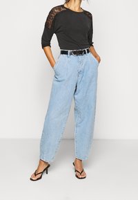 Top nero con maniche in pizzo, jeans a vita alta azzurri chiari e cintura nera. Sandali neri con tacco completano l'outfit.