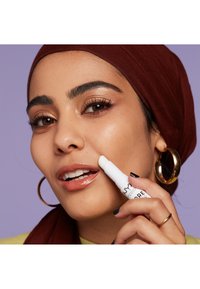 Bâtonnet cosmétique blanc tenu contre la joue, modèle avec des lèvres brillantes et un maquillage des yeux bronzé, portant un foulard bordeaux et des boucles d'oreilles en or.