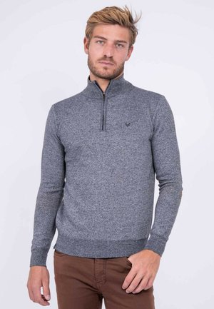 Homme aux cheveux blonds portant un pull gris à fermeture éclair partielle et un pantalon marron, debout devant un fond blanc uni.