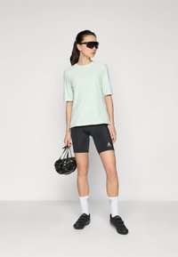 Camisa de manga curta verde claro, calções de ciclismo pretos e sapatos de ciclismo pretos. O modelo usa óculos de sol oversized pretos e segura um capacete.