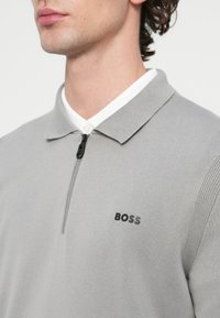 Szara koszulka polo z zamkiem błyskawicznym, prążkowaną teksturą, krótkim kołnierzykiem oraz czarnym logo "BOSS" po lewej stronie. Widoczny biały detal wewnętrznego kołnierzyka.