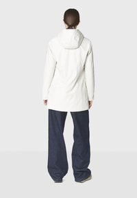 Ruskeat hiukset alhaalla poninhännällä oleva nainen valkoisessa hupullisessa takissa, leveissä tummansinisissä housuissa ja lenkkareissa, seisoo selin yksivärisellä taustalla.