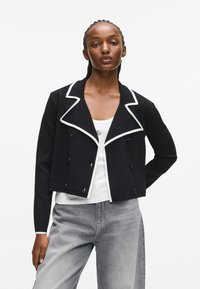 Czarna krótka kurtka z fakturowanego materiału, z białym kołnierzykiem, podwójnym zapięciem i czarnymi guzikami.