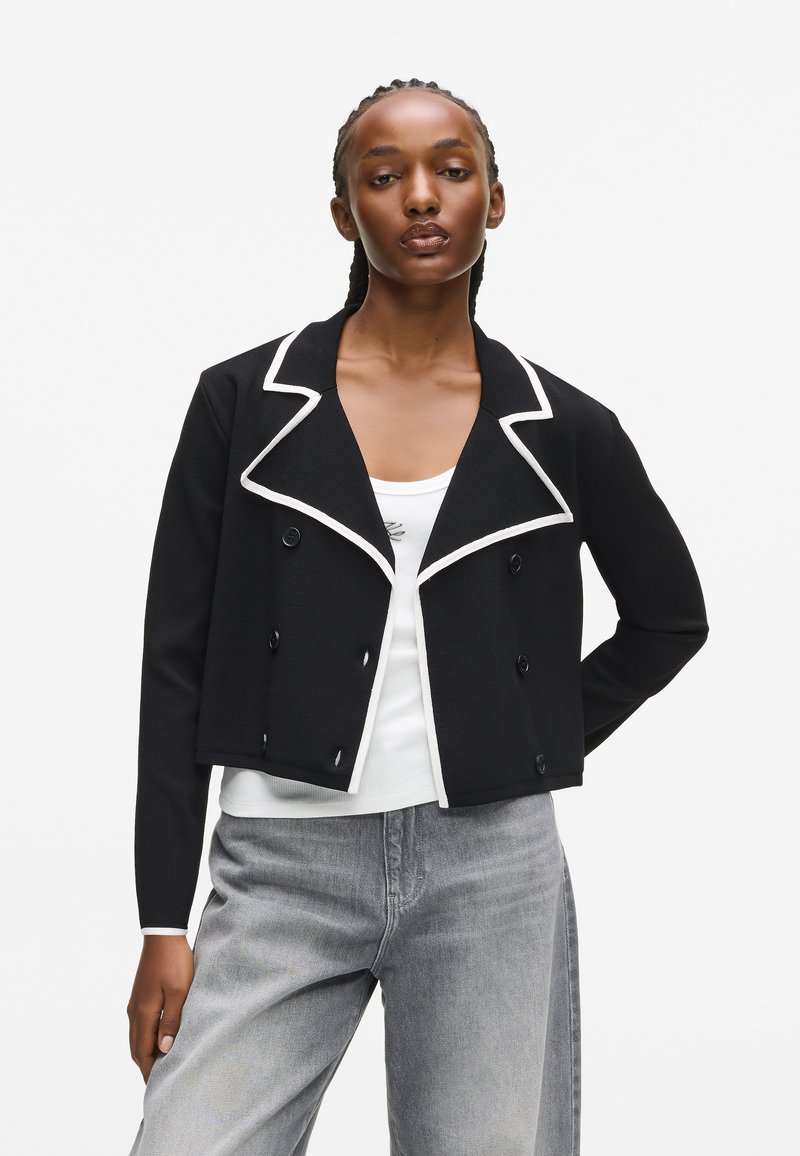 Czarna krótka kurtka z fakturowanego materiału, z białym kołnierzykiem, podwójnym zapięciem i czarnymi guzikami.