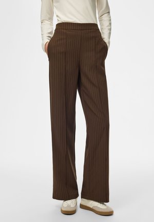 Personne portant un pantalon large marron à fines rayures et des baskets blanches avec des accents beiges, debout les mains dans les poches.