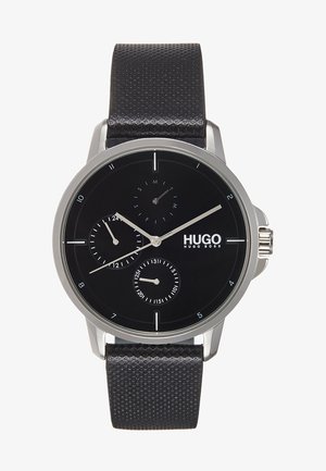 Montre analogique noire avec boîtier en acier inoxydable argenté, bracelet en cuir noir texturé, trois cadrans, et logo 'HUGO' bien en vue sur le cadran.