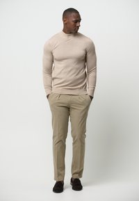Beige geribd langemouwen trui gecombineerd met licht khaki geplooide broek en donkerbruine loafers, wat zorgt voor een ontspannen, neutraal gekleed outfit.