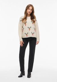 Pull en maille crème avec des motifs de rennes, nez rouges et pompons multicolores. Assorti à un pantalon en velours côtelé noir et des bottes noires à talons.