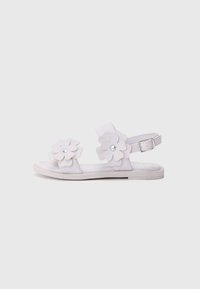 Hvit sandal med to blomsterdetaljer, hver med en sentral edelsten. Justerbar stropp og flat såle, laget av glatt syntetisk materiale.