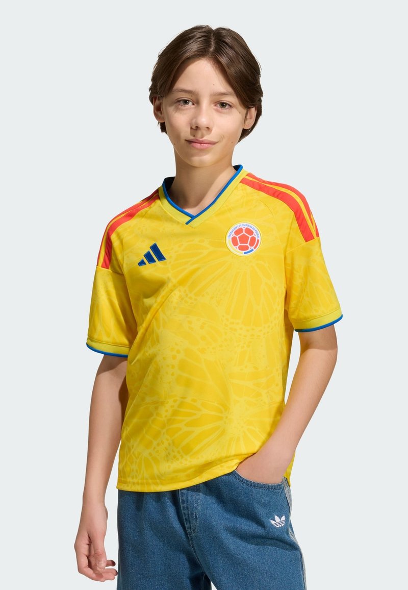 Maillot de football jaune à manches courtes, avec des accents bleus et des rayures rouges sur les épaules. Il présente un logo circulaire et un motif de papillon.