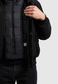 Czarna wielofunkcyjna kurtka z wewnętrzną warstwą na zamek błyskawiczny o pikowanym wzorze, zatrzaskami oraz małą metką z logo na podszewce.