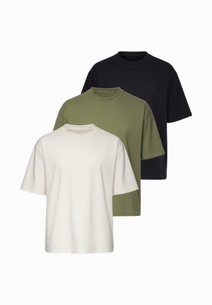 Jeune homme aux cheveux coiffés en arrière, portant un t-shirt beige oversize uni et un jean bleu clair, ajustant une montre-bracelet argentée.