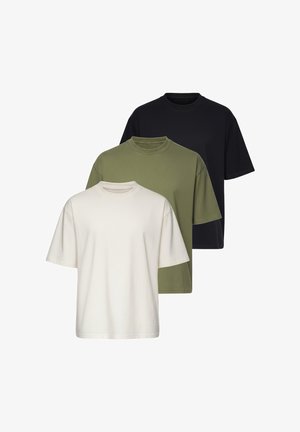 Jeune homme aux cheveux coiffés en arrière, portant un t-shirt beige oversize uni et un jean bleu clair, ajustant une montre-bracelet argentée.