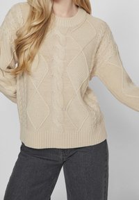 Beige stickad tröja med kabel- och diamantmönster, ribbat halsringning och nederkant, med avslappnad passform och strukturerad design. Matchad med mörka jeans.