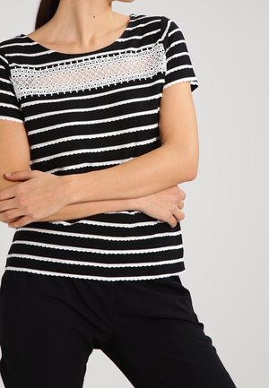 Mujer con una blusa de rayas blanco y negro de manga corta con detalles de encaje en el pecho, con los brazos cruzados, combinada con pantalones negros.